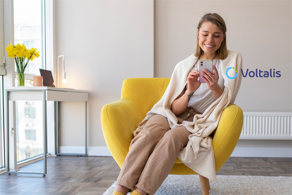 Femme souriante assise dans un fauteuil jaune, enroulée dans une couverture, utilisant son smartphone. À l’arrière-plan, un bureau avec un ordinateur portable et un vase de jonquilles jaunes près d’une fenêtre, avec le logo de l’entreprise Voltalis affiché sur le mur.